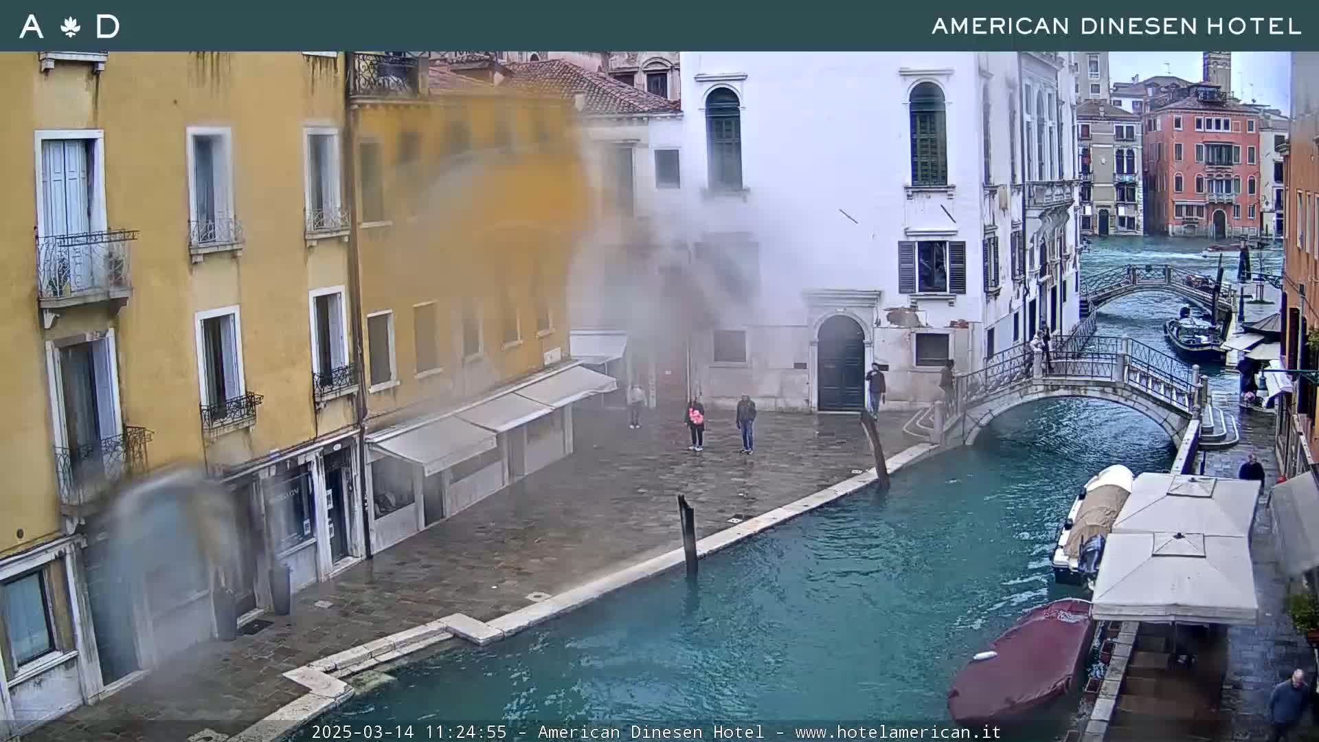 A misty scene of a canal in Venice, with several buildings, a bridge, and a few people on the waterfront.