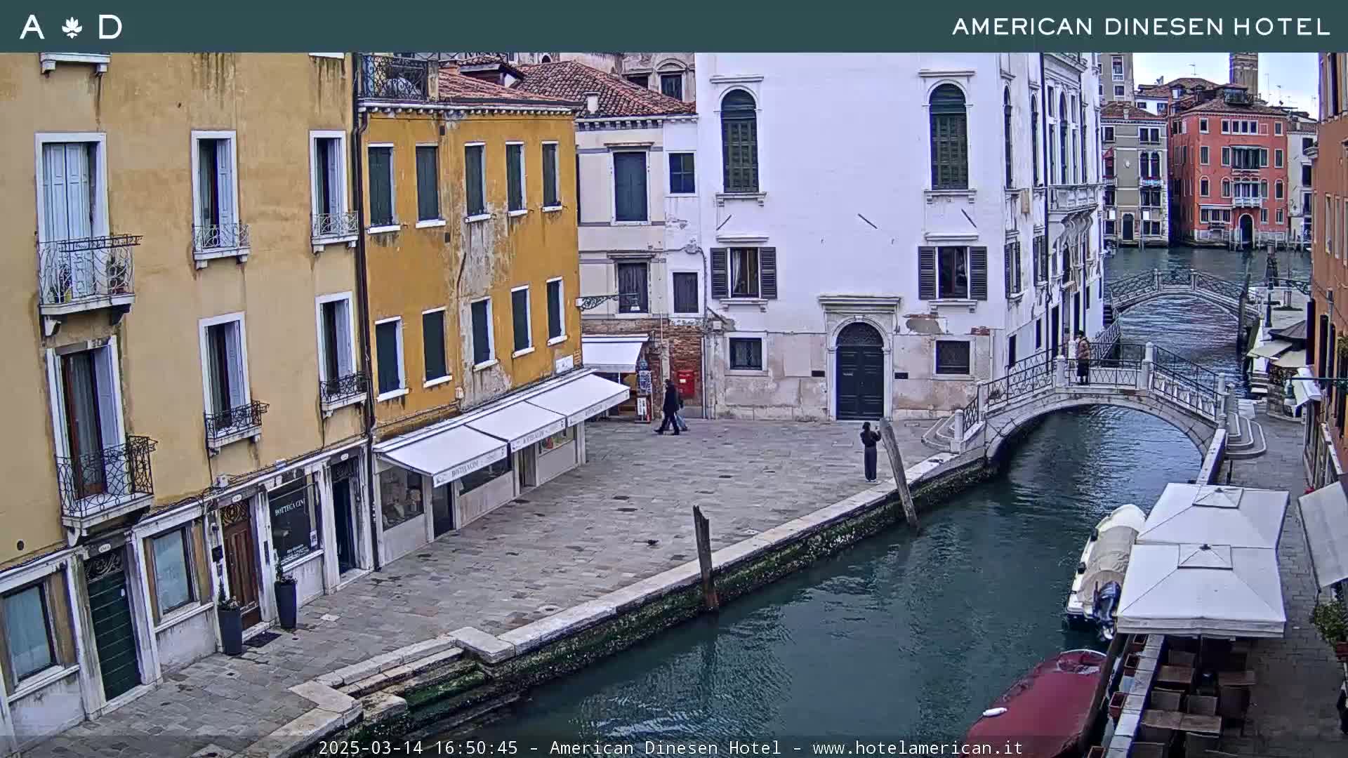 A view of a canal in Venice, Italy, shows several buildings along the waterway, a small bridge, and a few people walking along the canal side on an overcast day.
