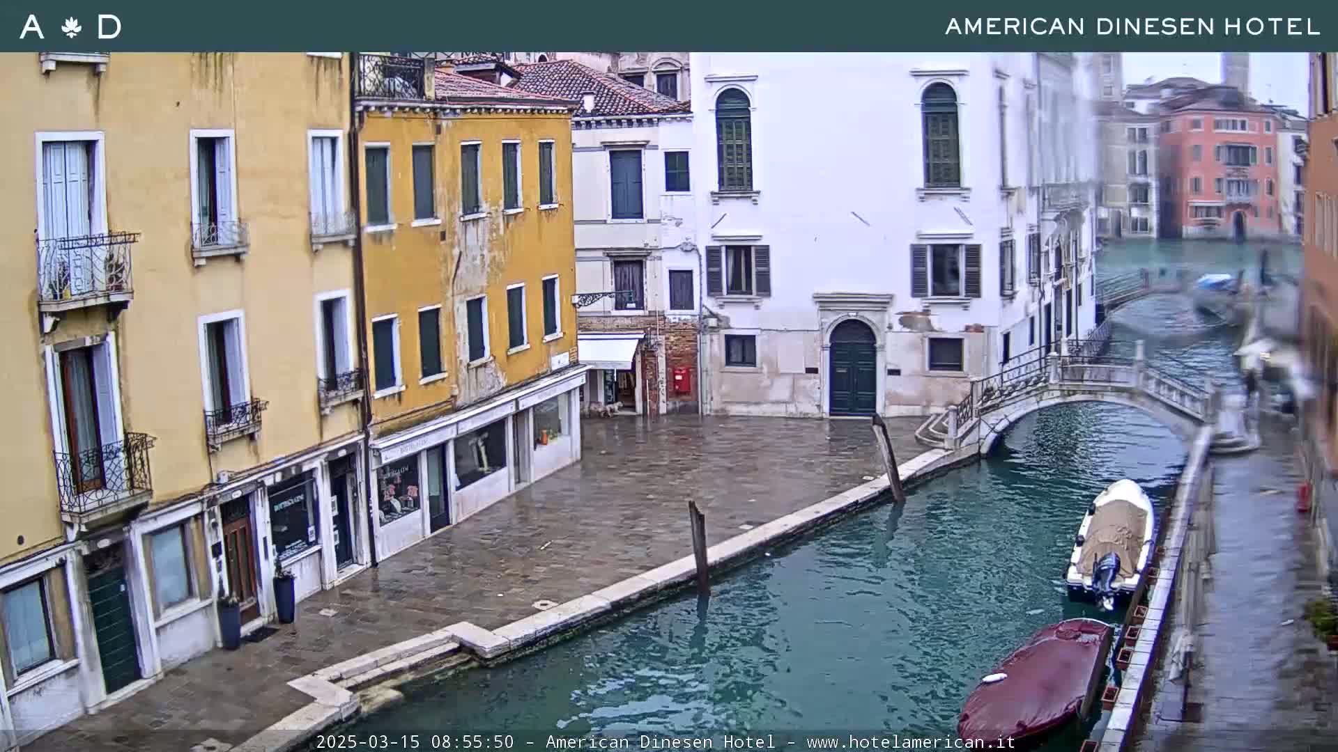 A view of a canal in Venice, Italy, on a cloudy day, shows several buildings lining the waterway, with boats moored along the canal's edge and a small bridge crossing it.