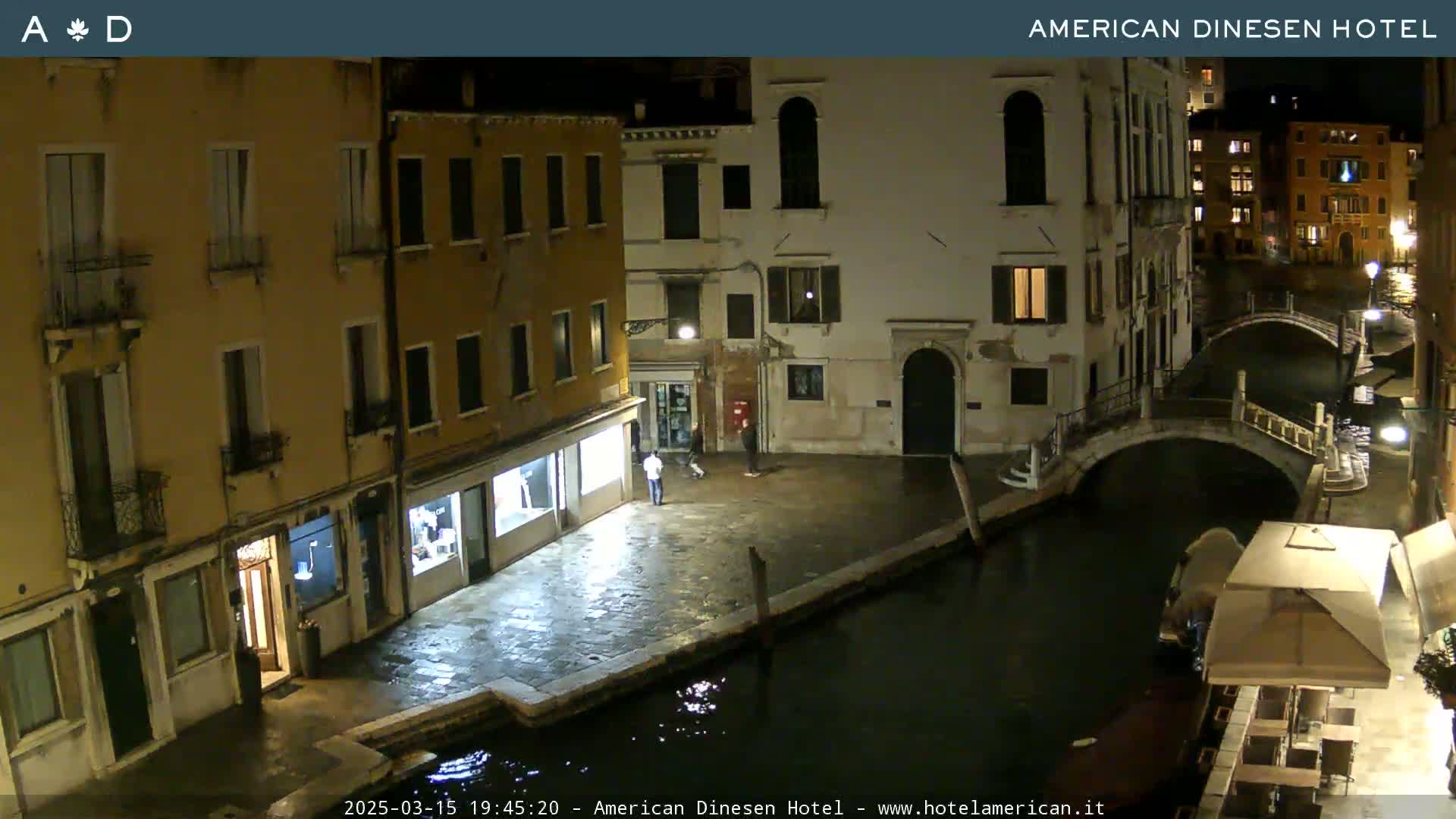 A nighttime view of a canal in Venice, showing several buildings lining the waterway, a small bridge, and a few people on the street.