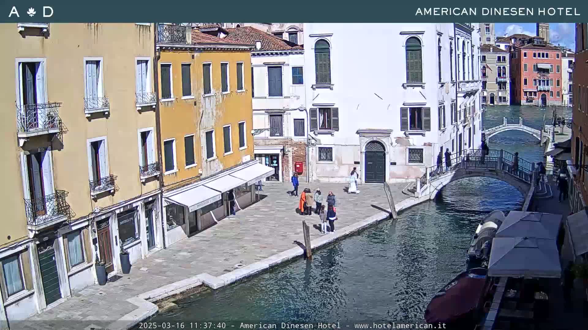 A sunny day in Venice shows several people walking near a canal with colorful buildings and a small bridge.
