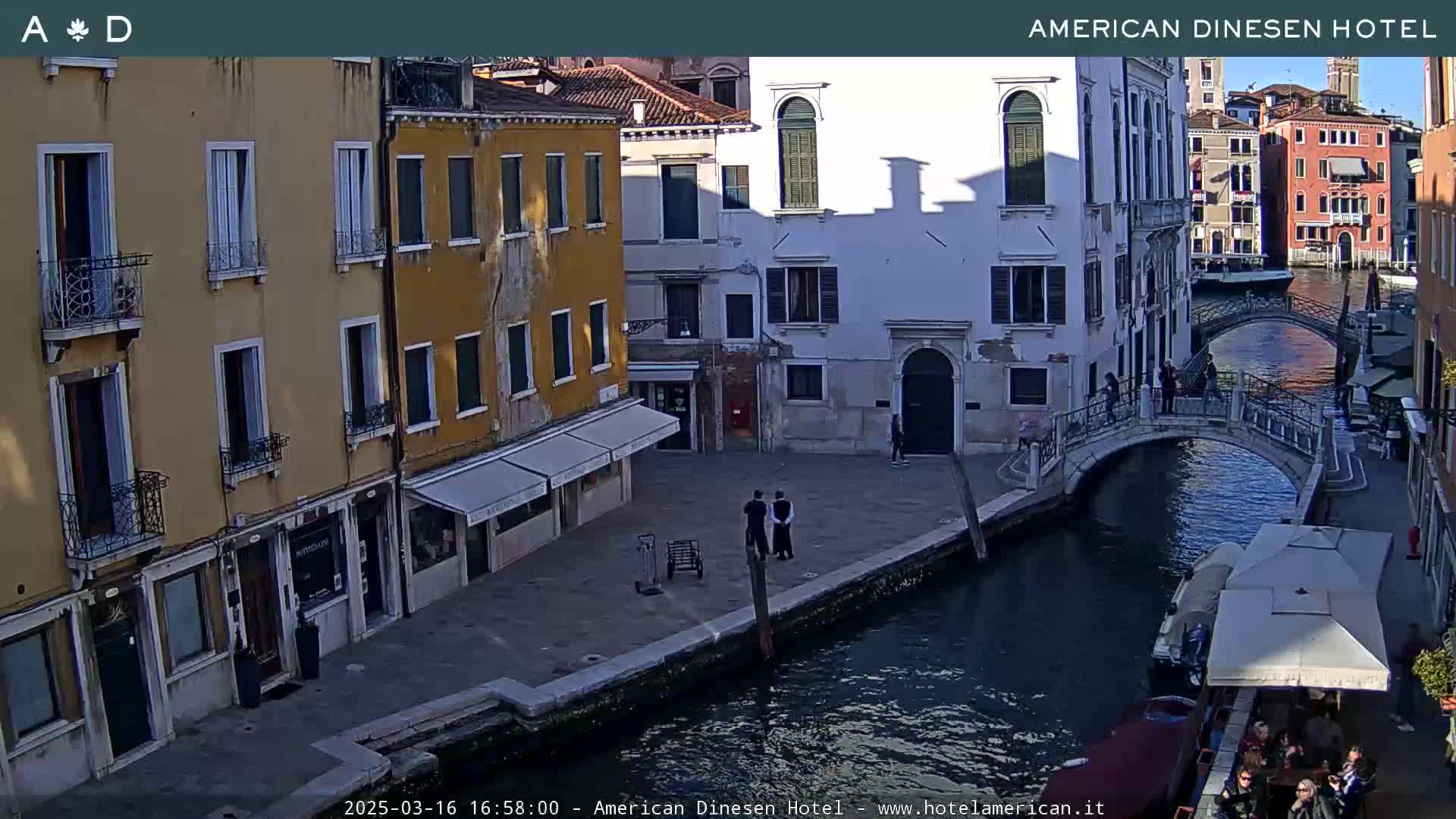 A sunny view of a canal in Venice, Italy, shows several buildings lining the waterway, a small bridge, and people walking along the canal and sitting at an outdoor café.