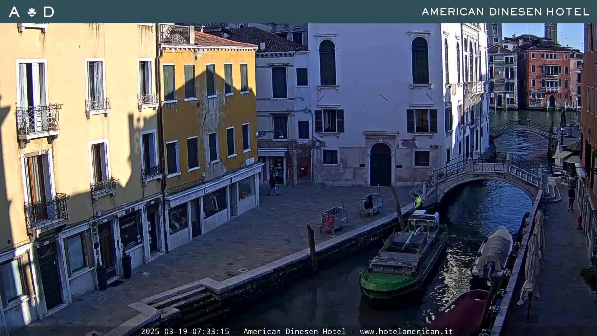 A sunny view of a canal in Venice, Italy, shows several buildings, a small bridge, and boats on the water.