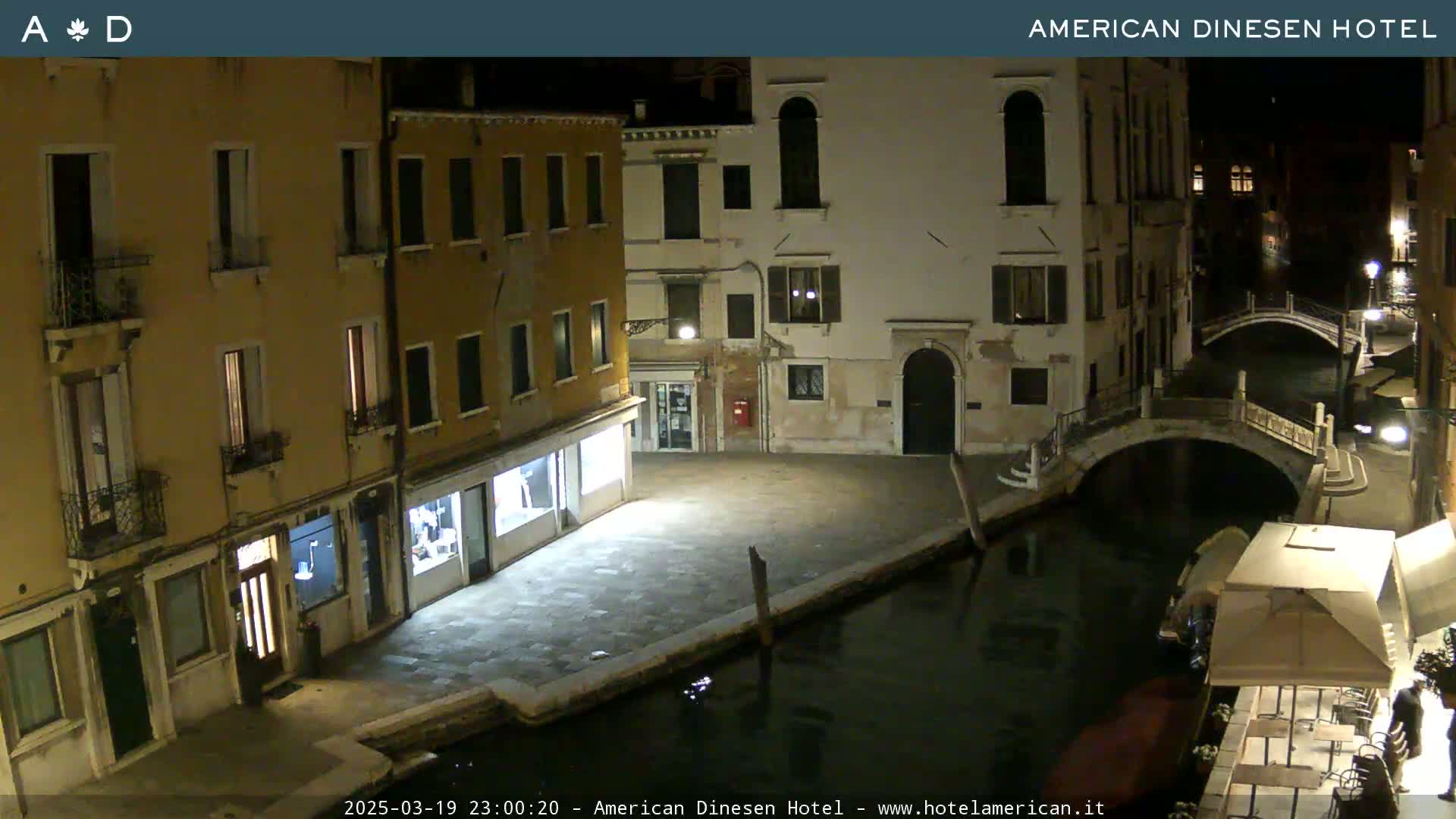 A nighttime view of a canal in Venice, Italy, shows several buildings lit along the waterway, with small bridges spanning the canal.