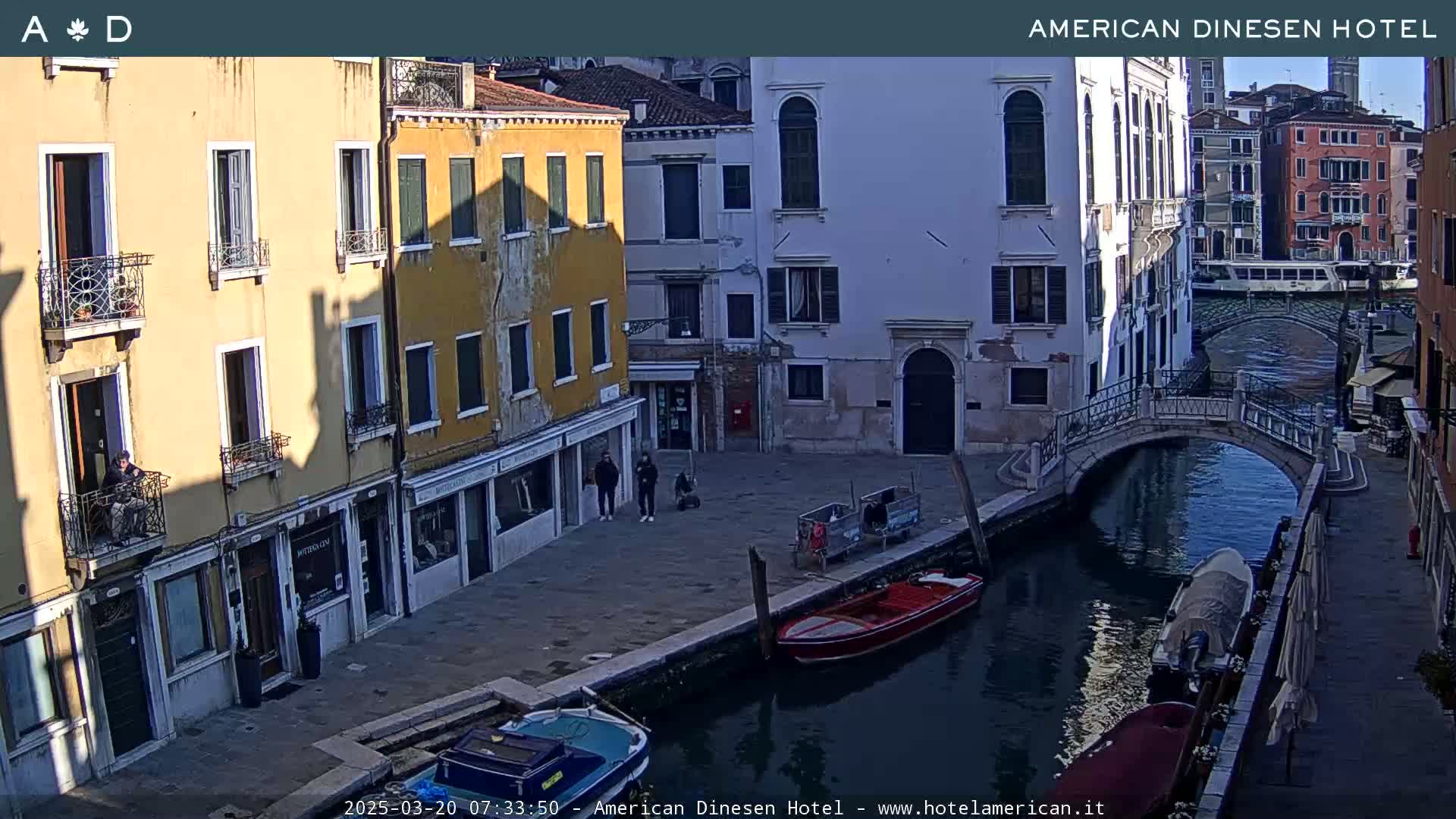 A sunny view of a canal in Venice, Italy, with colorful buildings lining the waterway and several boats moored alongside a stone bridge.