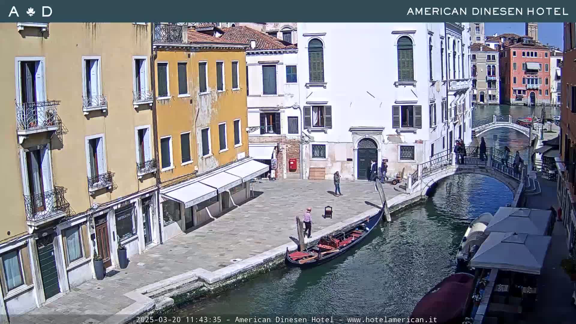 A sunny day in Venice shows a gondola on a canal next to buildings and a small bridge with several people walking around.