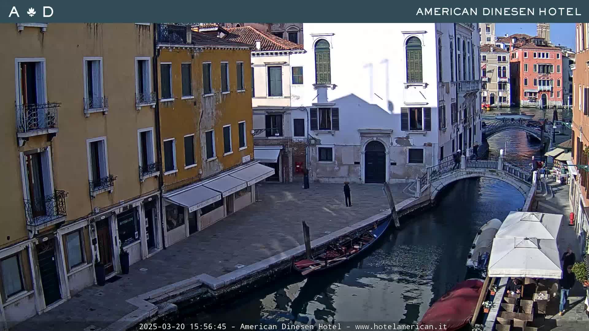 A sunny Venetian canal scene shows colorful buildings, a gondola, a bridge, and several people walking along the waterway.