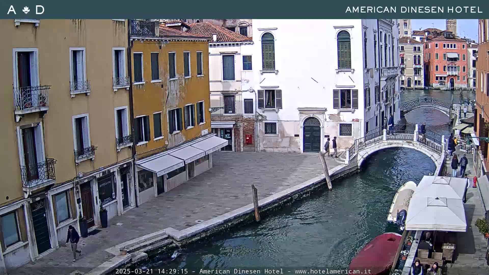A sunny view of a canal in Venice, Italy, shows several buildings lining the waterway, a small bridge crossing the canal, and a few pedestrians.