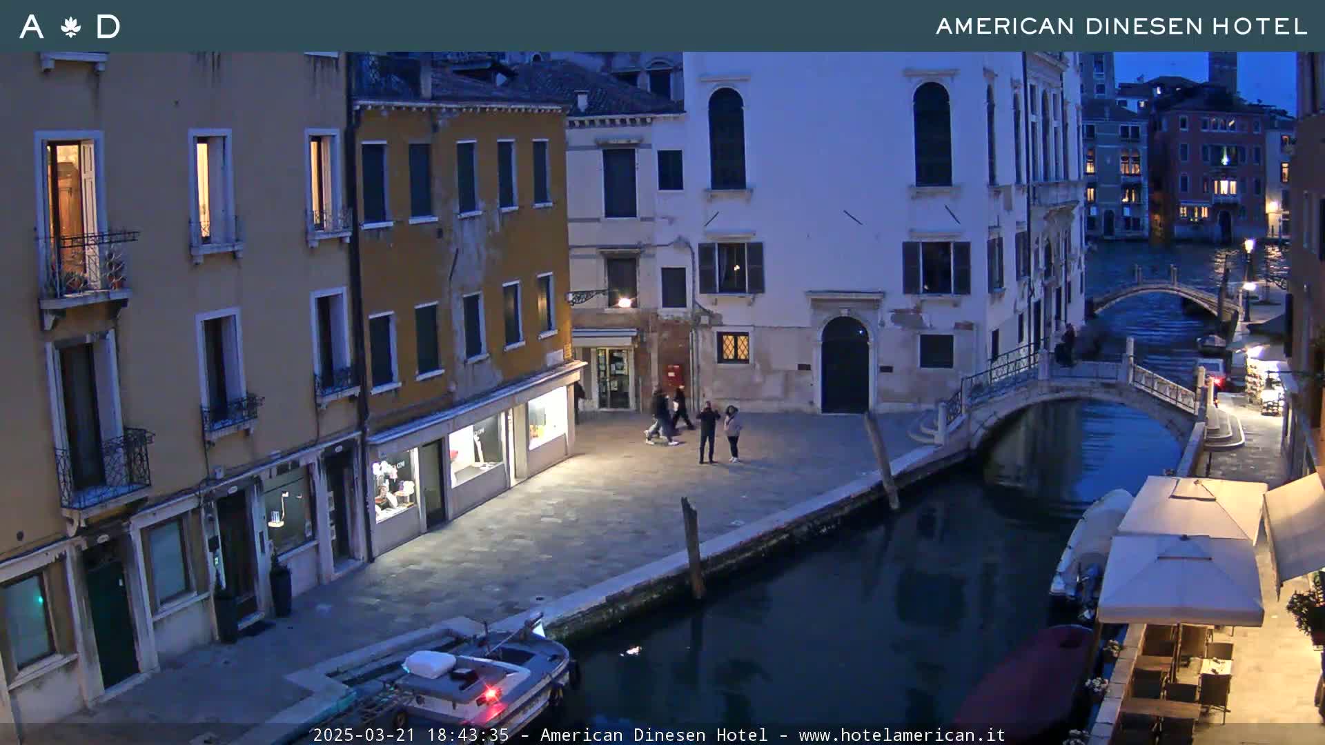 A twilight view of a canal in Venice, showing several buildings and a few pedestrians along the waterway, with a small boat moored nearby.