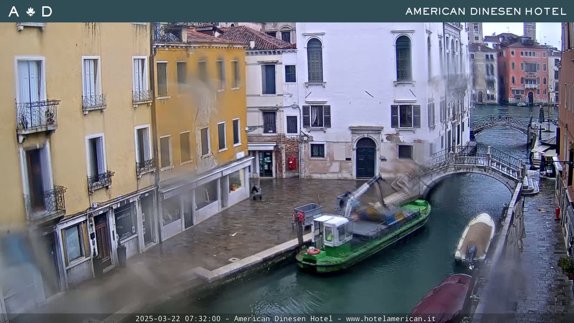 A rain-slicked canal scene in Venice shows several buildings lining the waterway, a small green barge, and a pedestrian bridge.