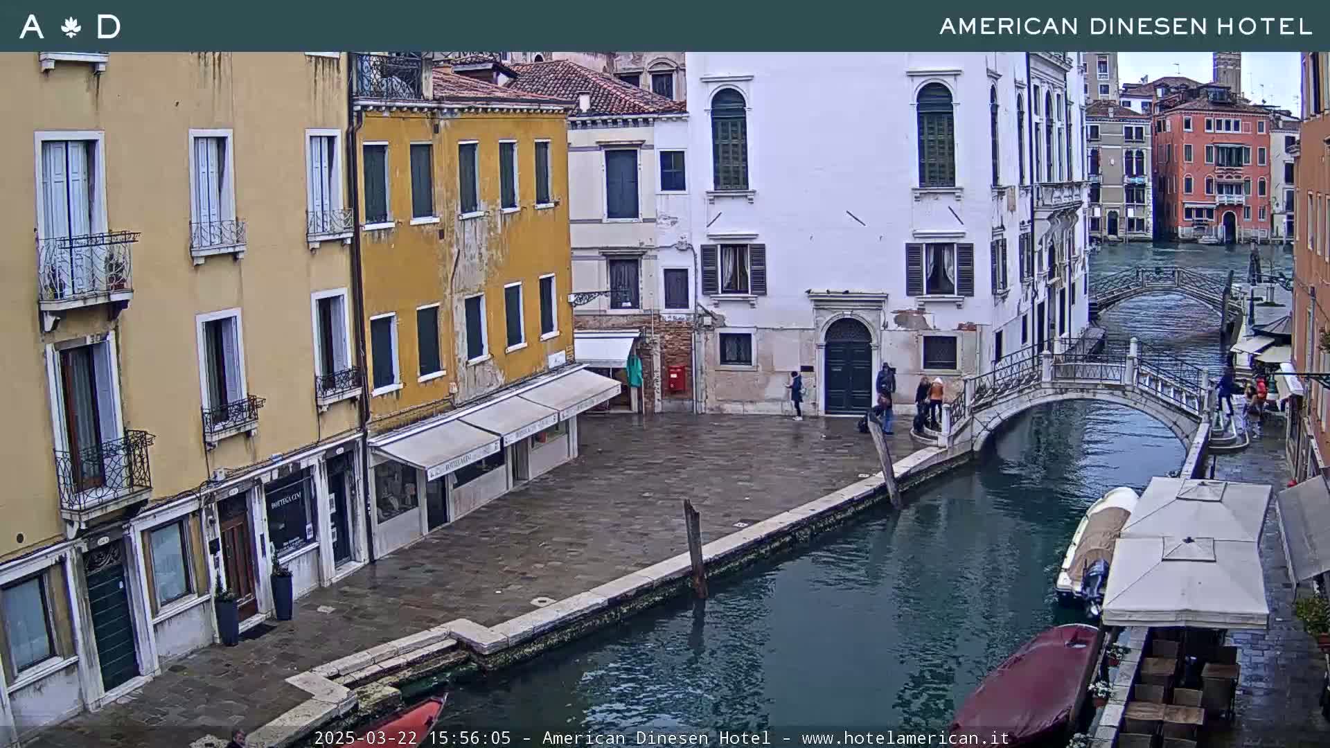 A canal in Venice, Italy, is shown on an overcast day; several people stand on a stone embankment next to buildings and a small bridge.