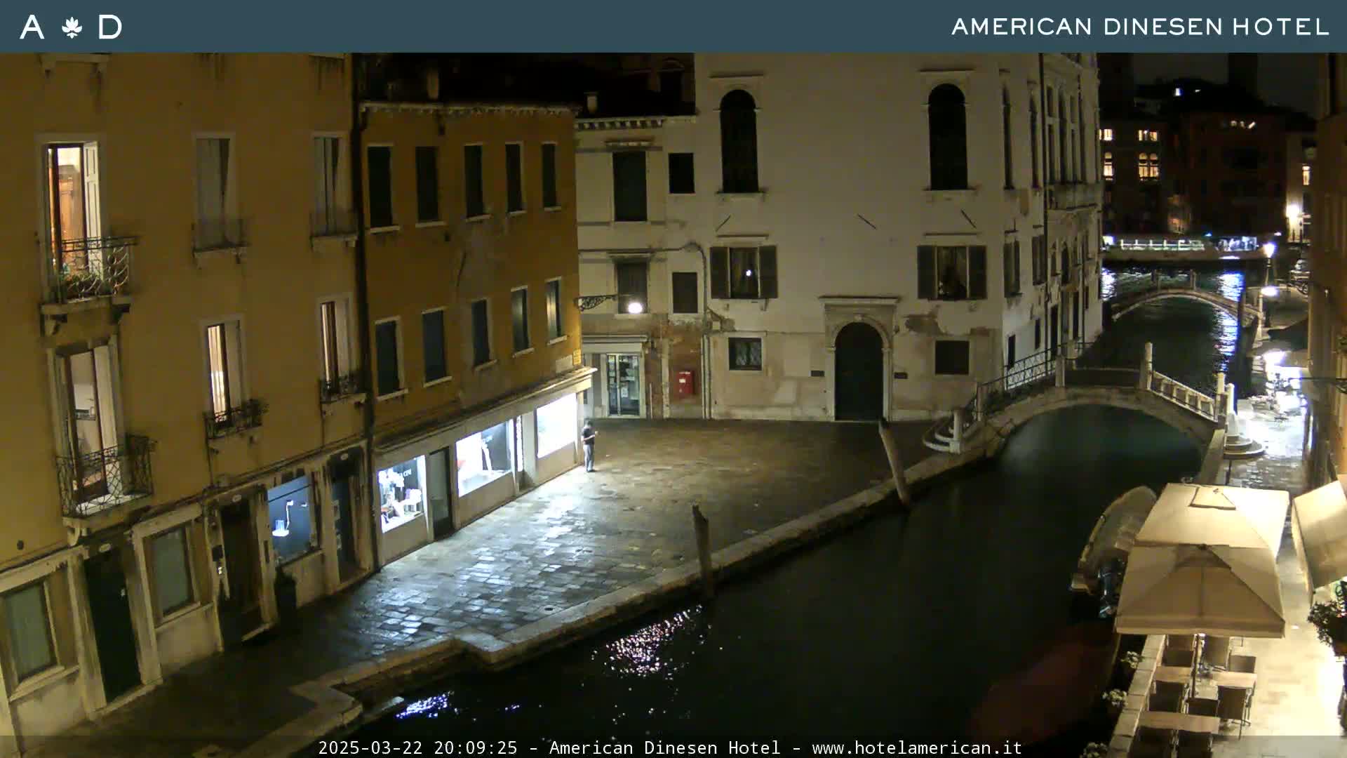A nighttime view of a canal in Venice, Italy, with stone buildings lining the waterway and a bridge visible in the distance.