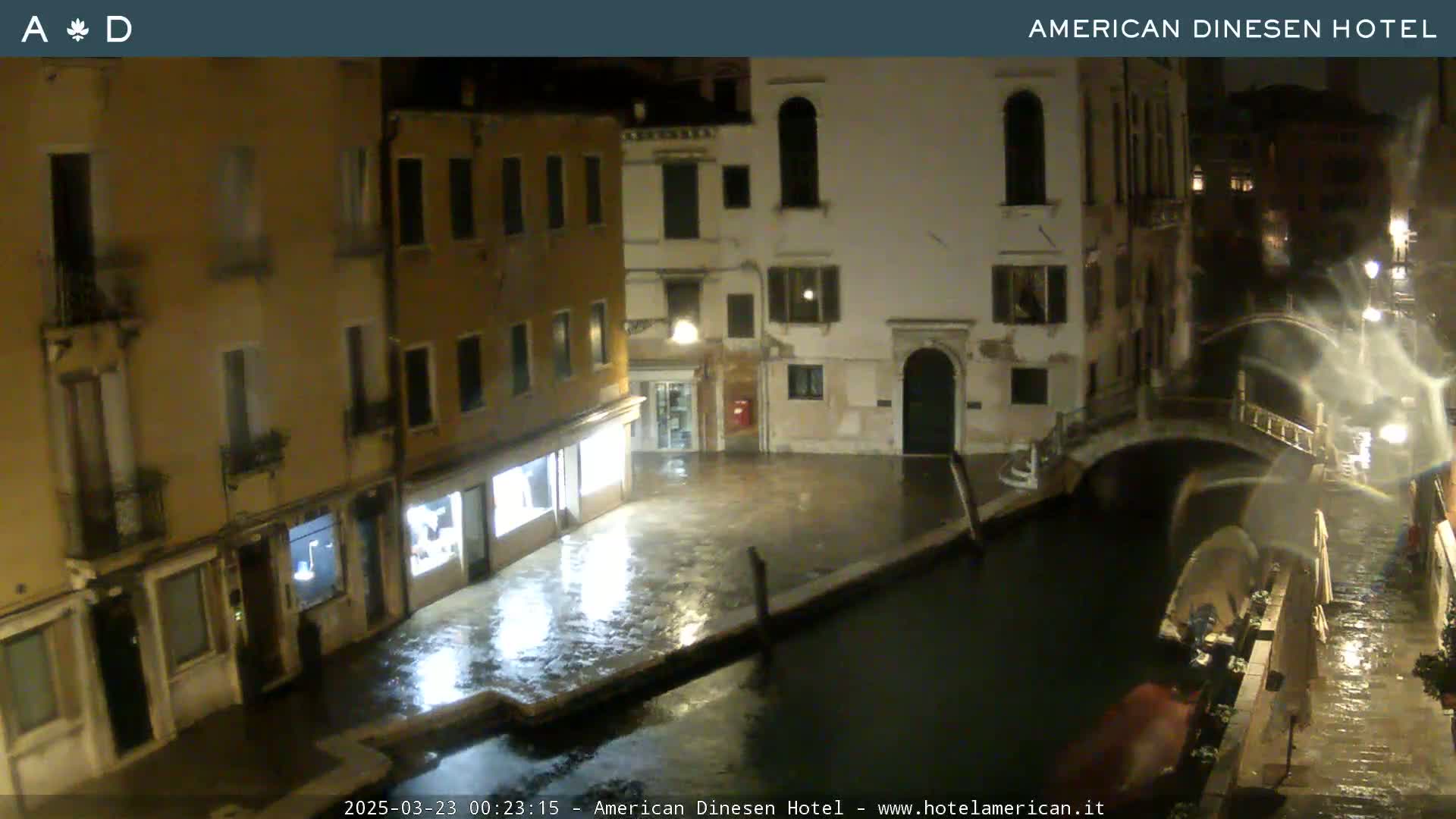 A nighttime view of a canal in Venice, Italy, showing wet cobblestone streets and buildings under rainy conditions.