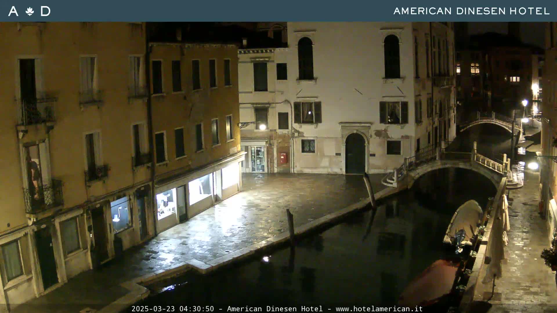 A nighttime view of a canal in Venice, showing several buildings along the waterway and a small bridge, with a boat moored alongside the canal.