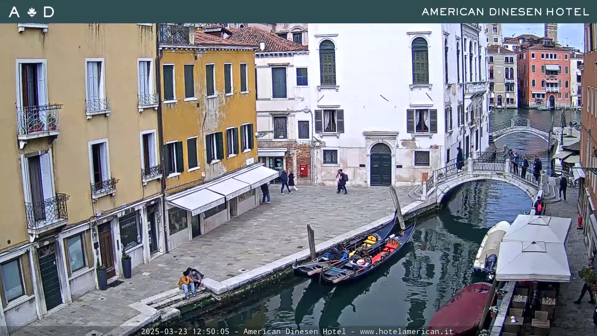 A gondola is moored along a canal in Venice, Italy, under a partly cloudy sky, with pedestrians on a nearby bridge and several buildings lining the waterway.
