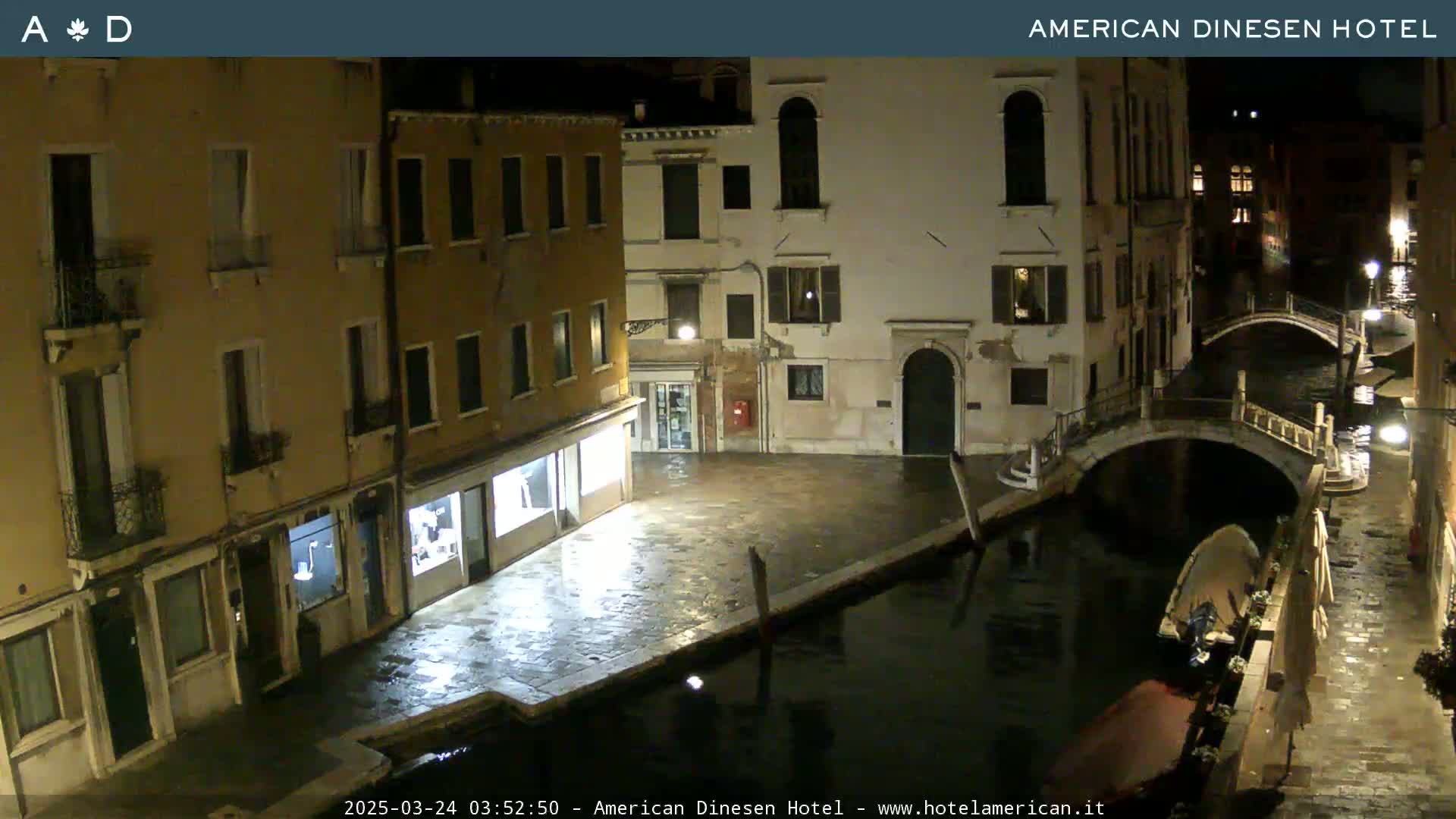 A nighttime view of a canal in Venice, Italy, showing several buildings along the waterway and a small bridge, with wet pavement visible.