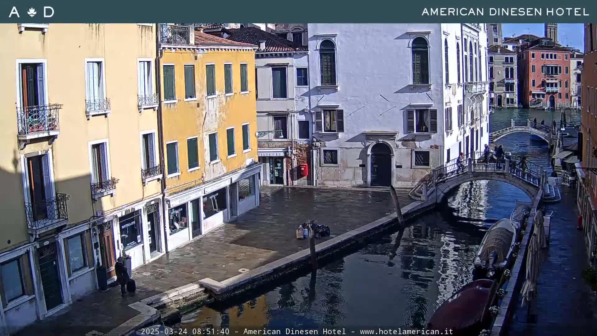 A sunny view of a canal in Venice, Italy, with several buildings lining the waterway and a small bridge connecting the two sides.