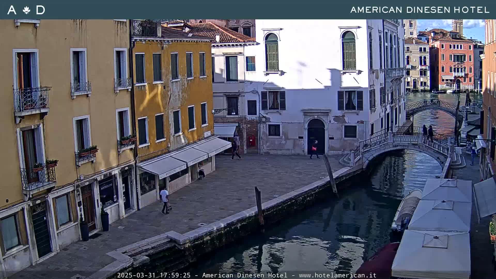 A sunny view of a canal in Venice, Italy, shows several buildings, a bridge, and a few pedestrians.