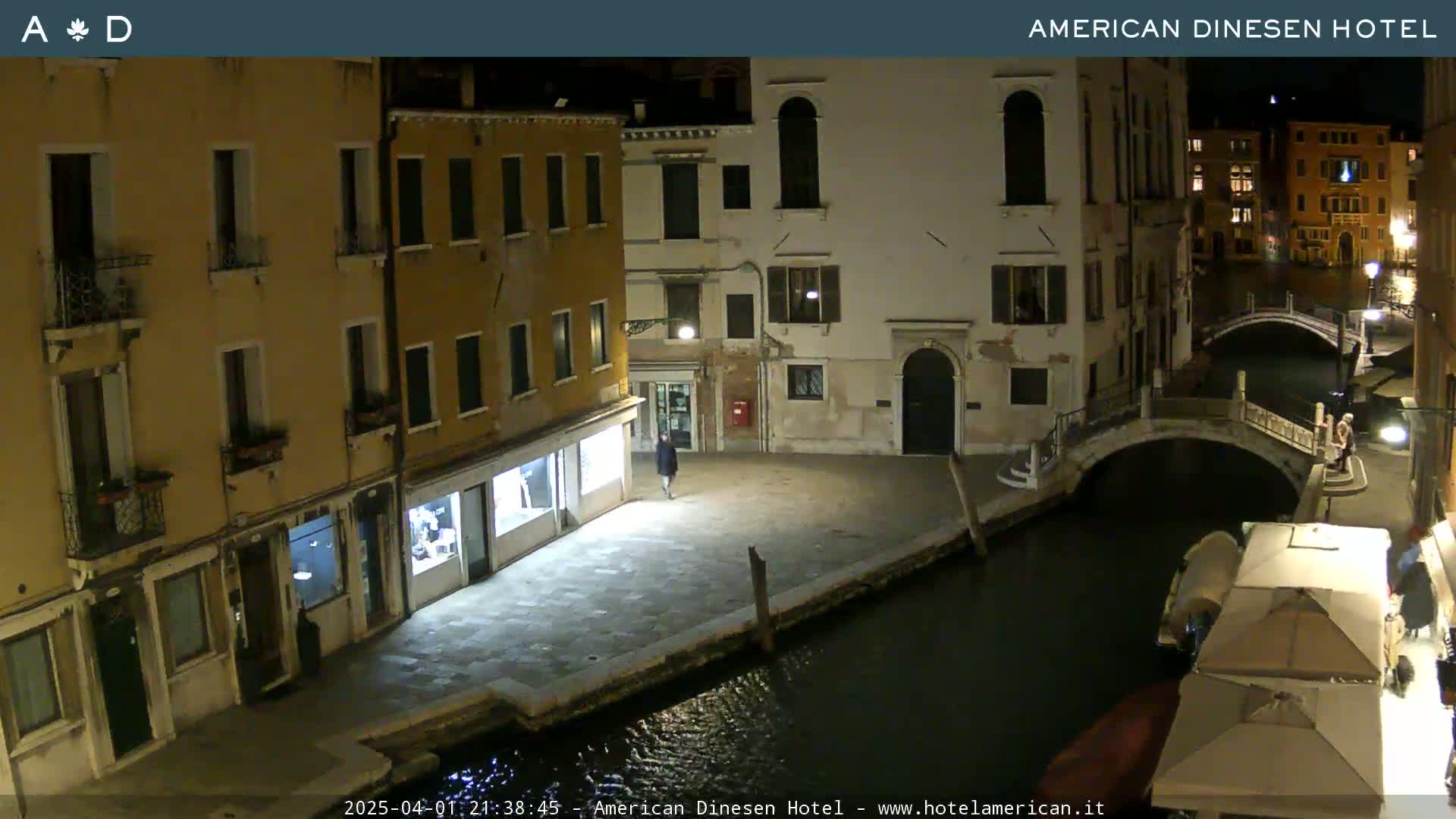 A nighttime view of a canal in Venice, Italy, shows several buildings lit along the waterway with a small bridge and a few people visible.