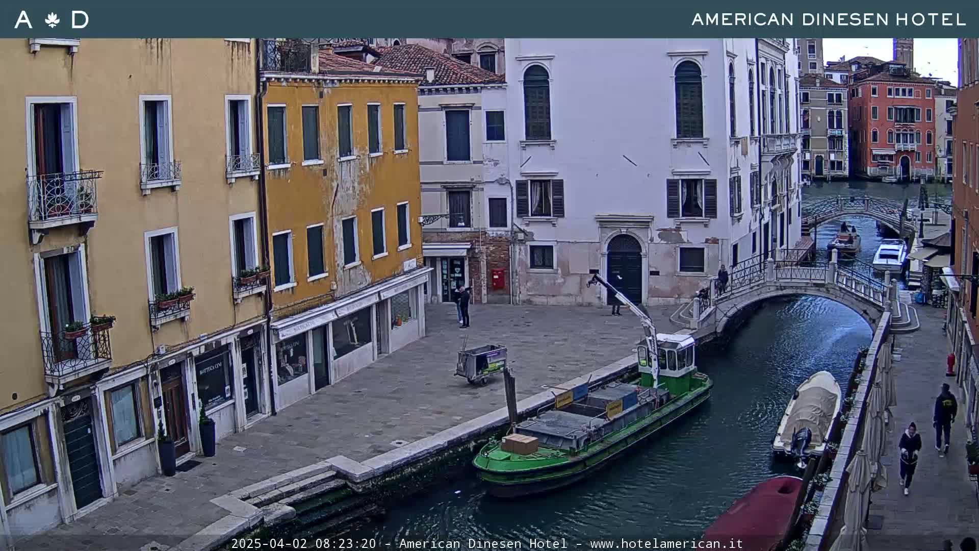 A canal in Venice, Italy, with several buildings lining its banks, a barge on the water, and a few pedestrians on the street.