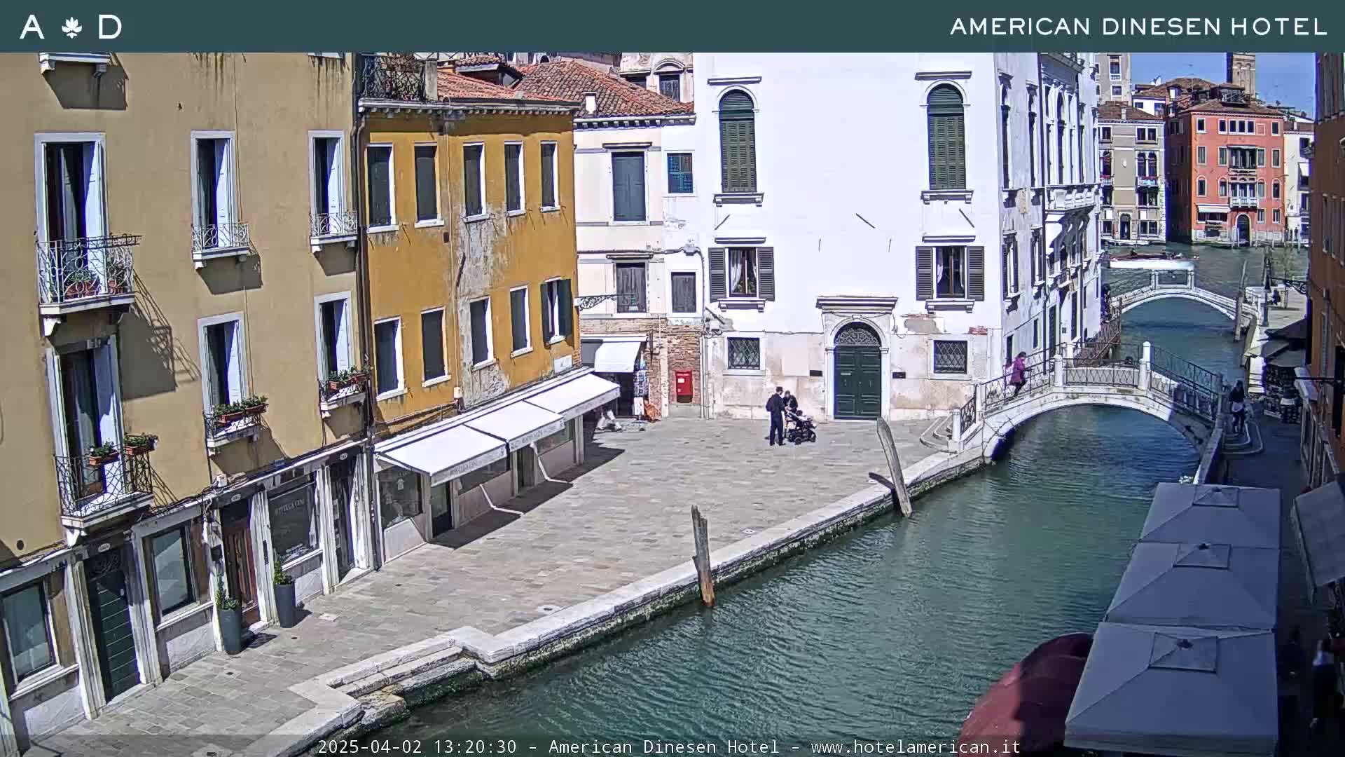 A sunny day in Venice shows a canal with two bridges, lined with colorful buildings and a few people walking near shops with awnings.