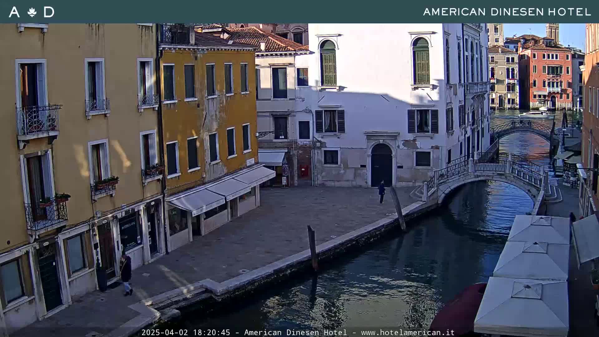 A sunny view of a canal in Venice, Italy, shows several buildings along the waterway, a small bridge, and a few people walking.