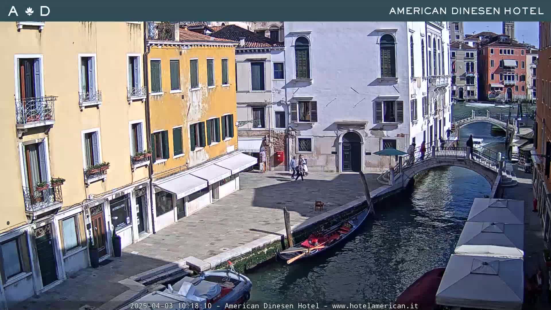 A sunny day in Venice shows a canal with a gondola, a small bridge, and several buildings lining the waterway.