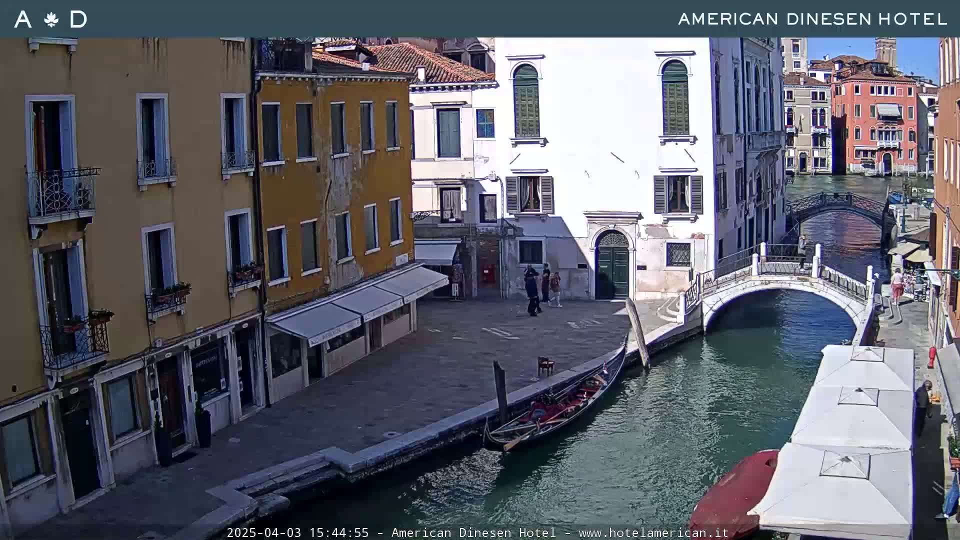 A sunny view of a canal in Venice, Italy, shows several buildings, a gondola, a small bridge, and some people walking along the canal.