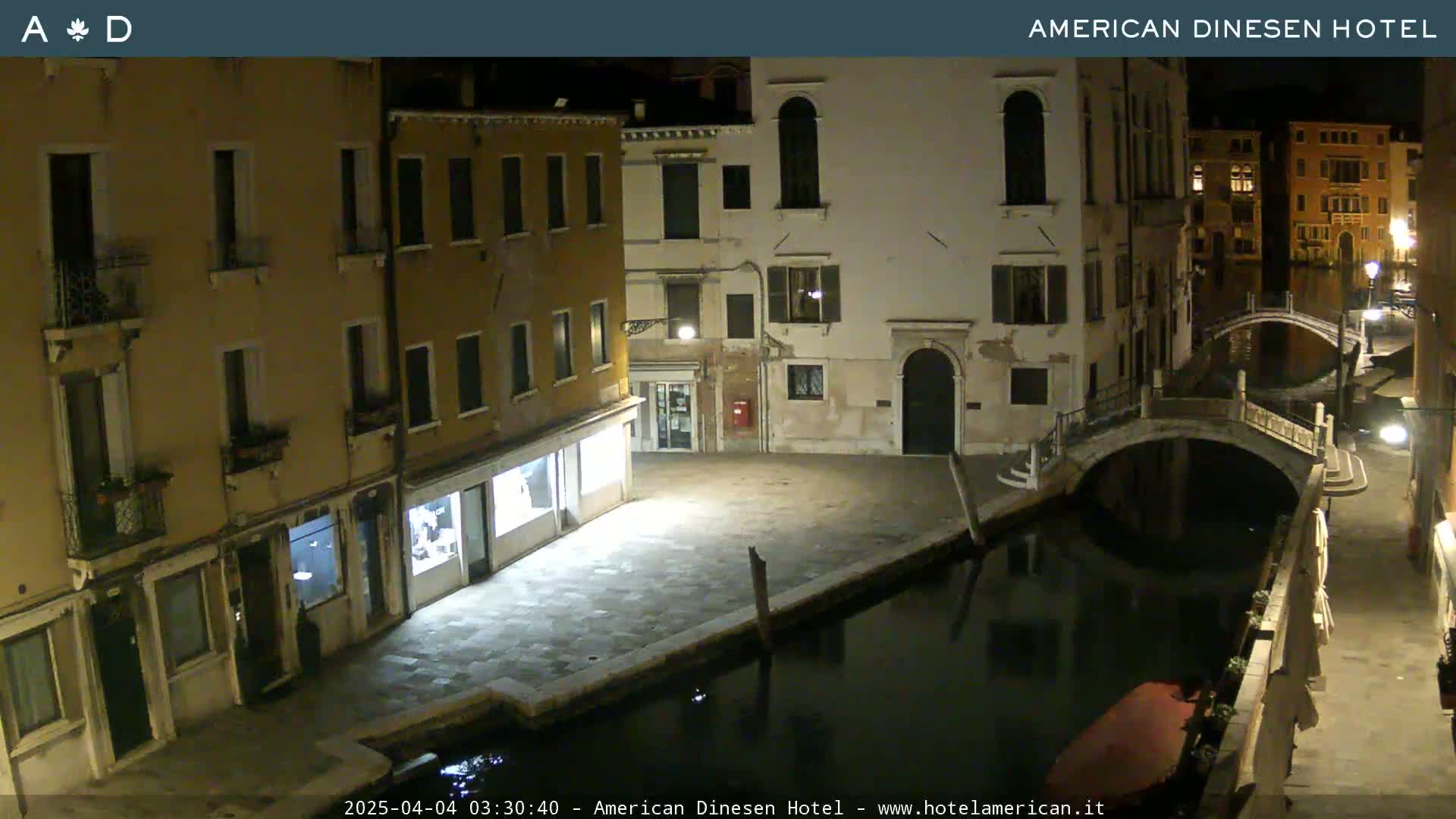 At night, a canal in Venice flows between stone buildings and under a small bridge, with a boat moored nearby.