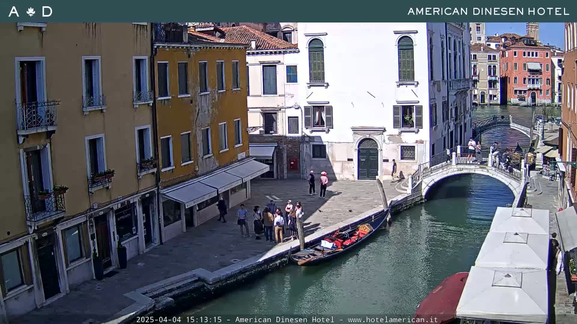 A sunny day in Venice shows a canal with a gondola, a small bridge, and several people walking along the waterway and buildings.