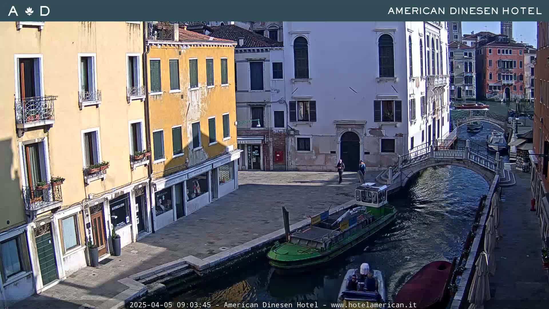 A sunny view of a canal in Venice, Italy, shows several buildings lining the waterway, a small bridge, and several boats on the canal.