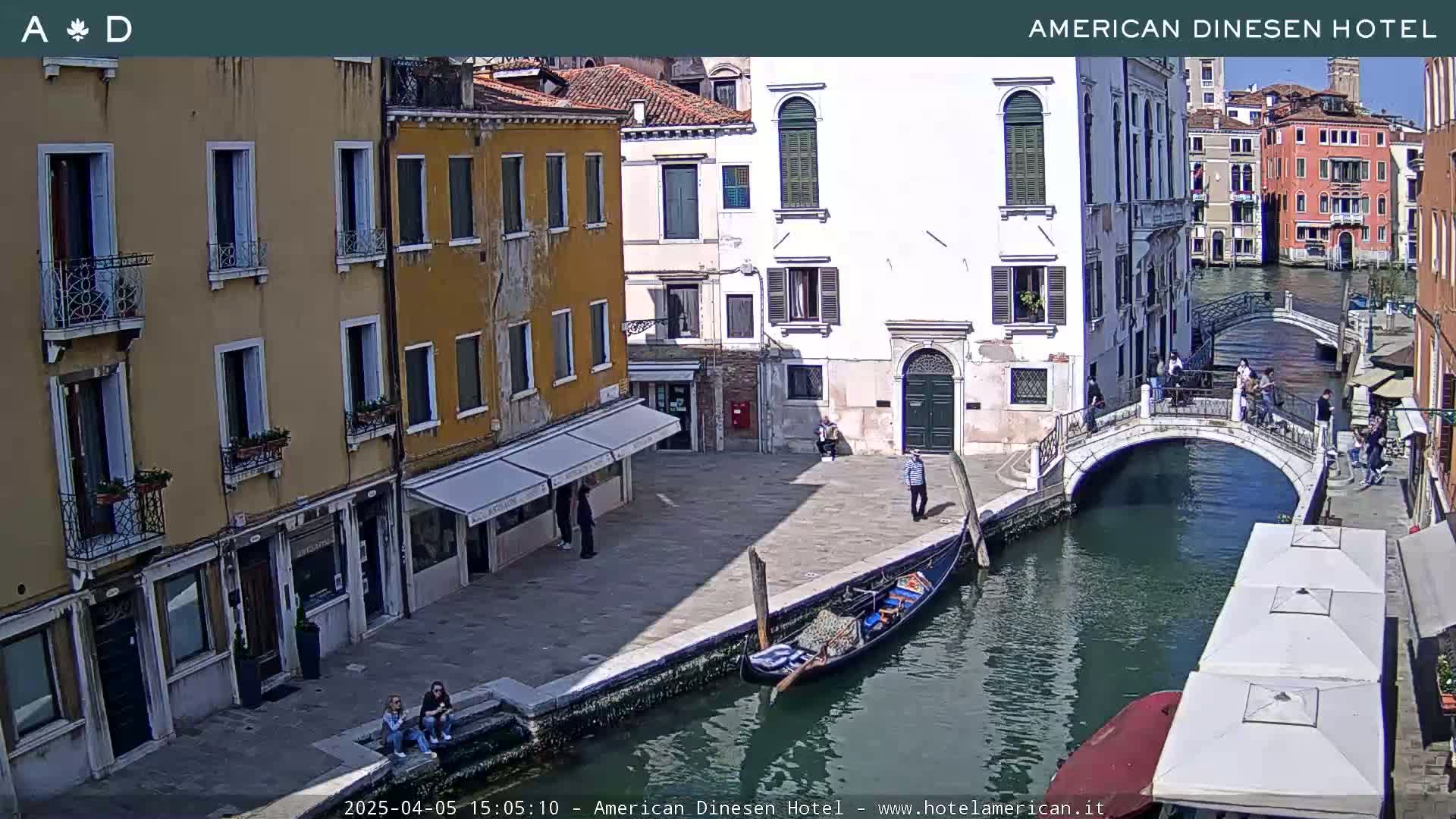 A sunny view of a canal in Venice, Italy, shows several buildings, a gondola, a bridge with people on it, and a few people walking along the canal.