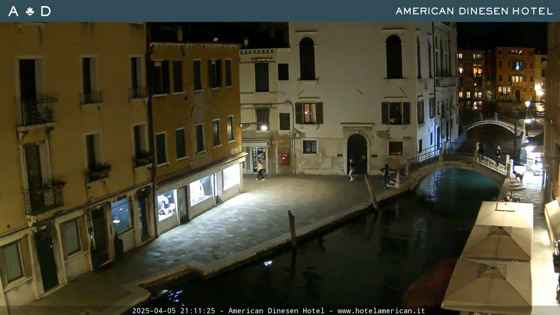 A nighttime view of a canal in Venice, with several buildings lining the waterway, and a few pedestrians visible.