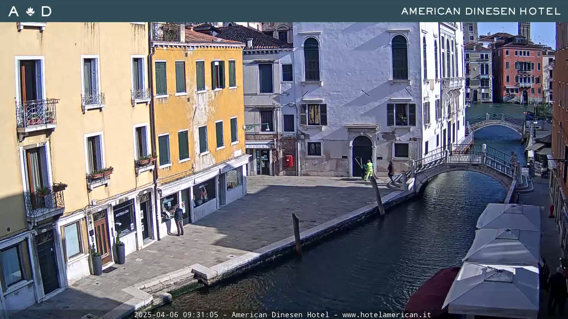 A sunny day in Venice shows a canal with a bridge, colorful buildings lining the waterway, and a few pedestrians.