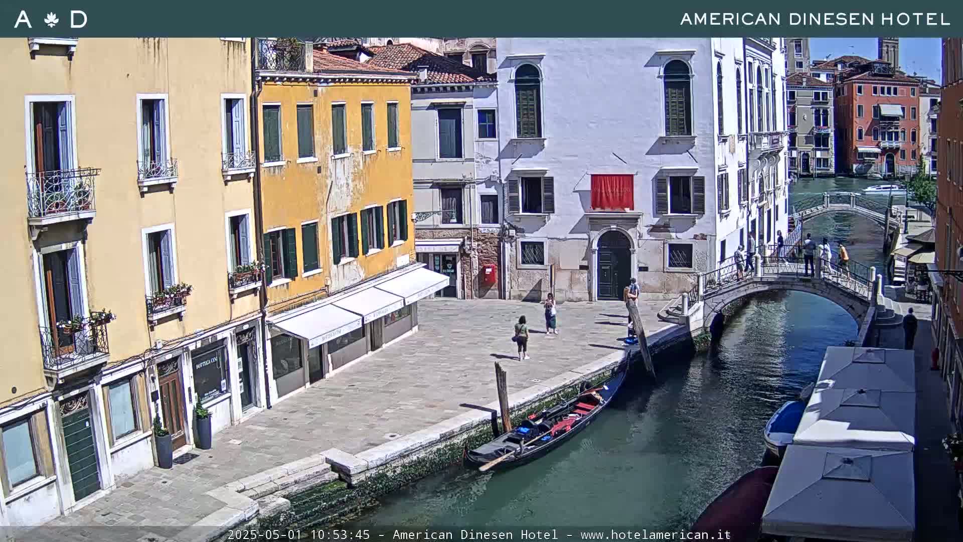 A sunny Venetian canal scene shows colorful buildings, a gondola, a footbridge, and several people strolling along the waterfront.