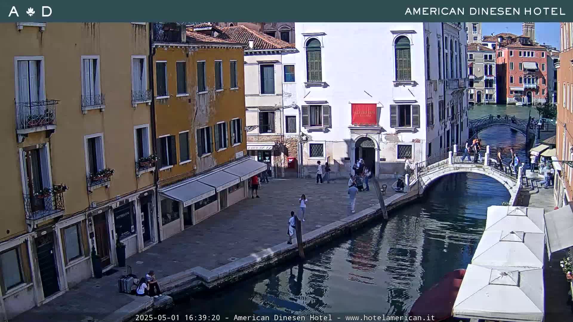 A sunny day in Venice shows a canal with a small bridge, several pedestrians, and buildings lining the waterway.