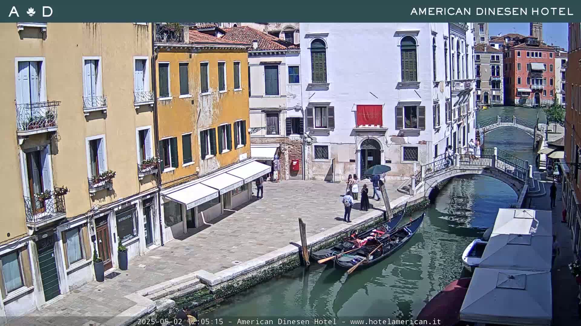 A sunny day in Venice, showing a canal with gondolas, a bridge, and several buildings lining the waterway, with people walking near a hotel.
