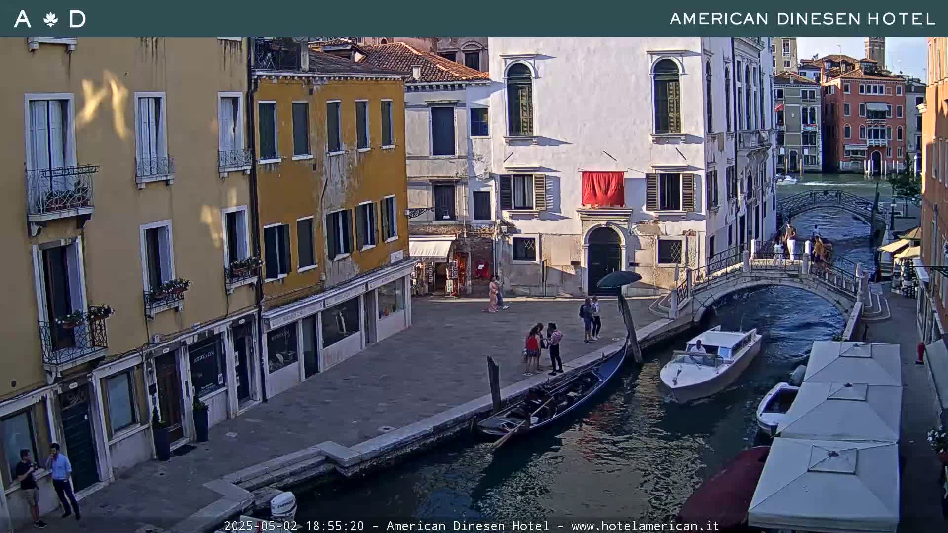 A sunny Venetian canal scene shows colorful buildings lining the waterway, with a gondola and motorboat on the water and several pedestrians on a bridge and the adjacent walkways.