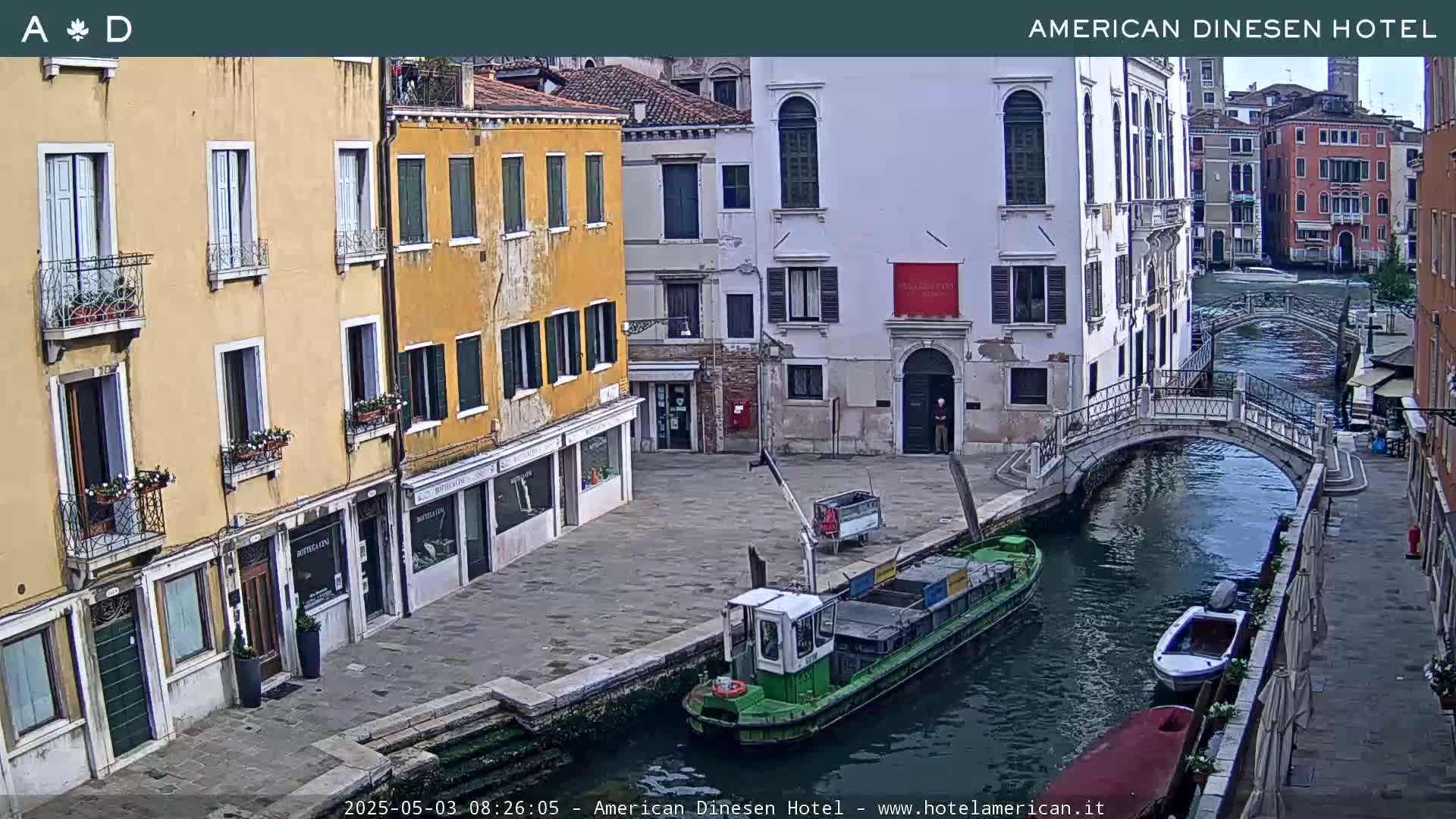 A canal in Venice, Italy, on a sunny day, shows several buildings lining the waterway, with a small green barge and other boats moored along the canal.