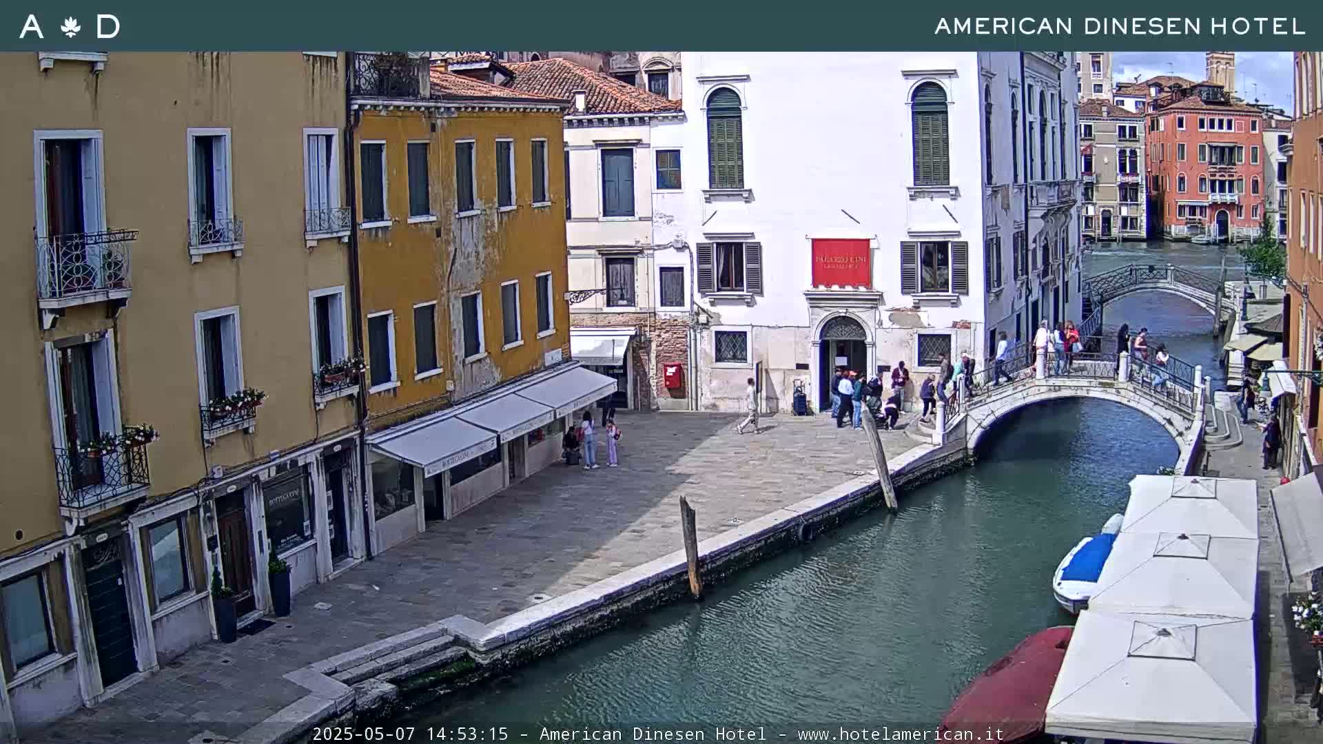 A sunny view of a canal in Venice, Italy, shows buildings lining the waterway, a footbridge spanning the canal, and several people walking along the canal and bridge.