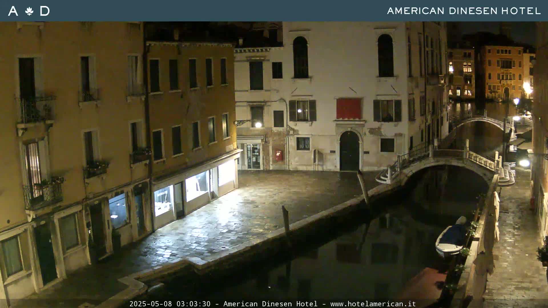 A nighttime view of a canal in Venice, Italy, showing buildings lining the waterway and a small boat moored alongside a bridge.
