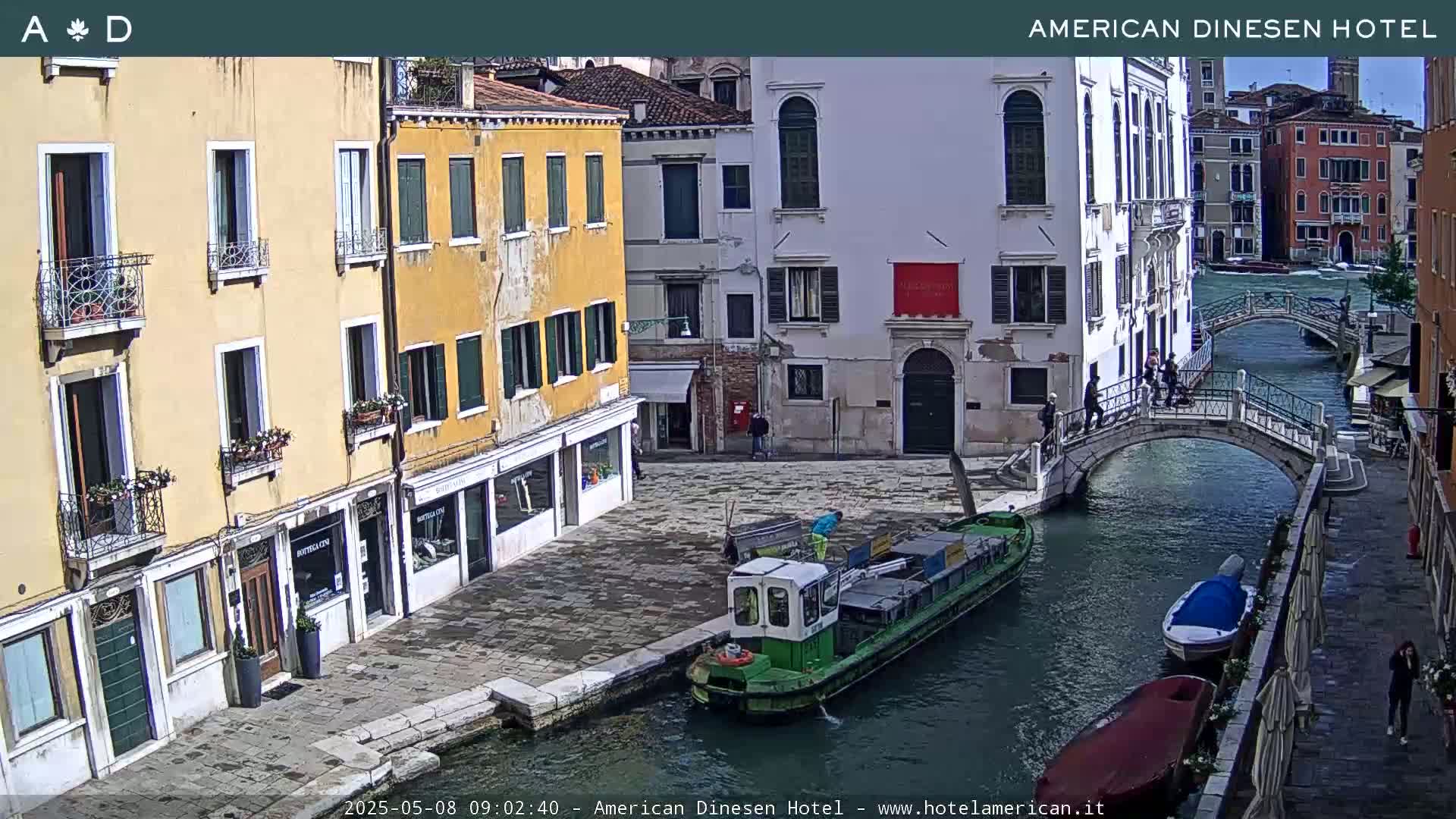 A sunny view of a canal in Venice, Italy, shows several buildings lining the waterway, with a small barge and other boats moored along the canal, and a pedestrian bridge crossing it.