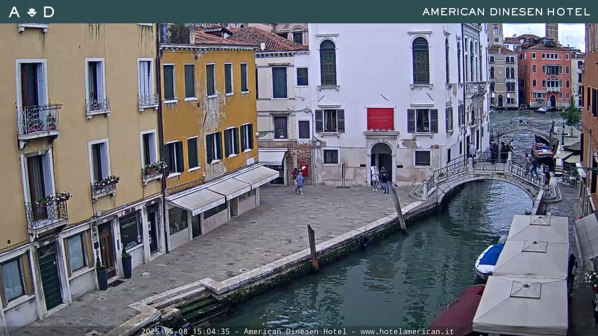 A canal in Venice, Italy, with several buildings lining its banks, a small bridge spanning the waterway, and a few people walking along the pathways.
