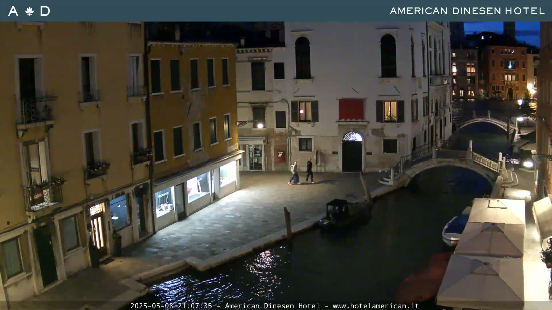A nighttime view of a canal in Venice, showing several buildings and a small bridge, with a couple walking on a stone path next to the water.