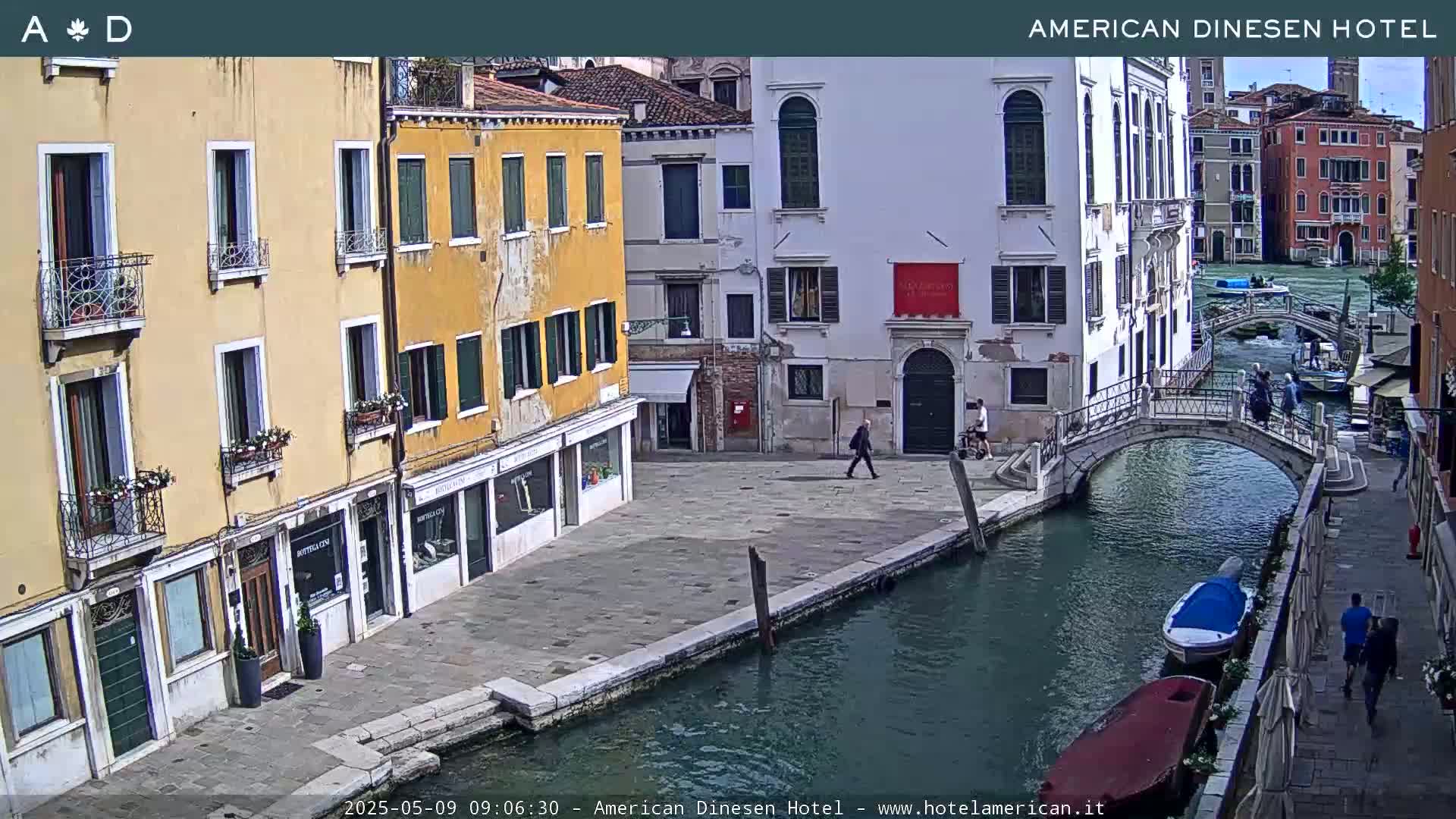 A sunny view of a canal in Venice, Italy, shows several buildings lining the waterway, a small bridge, and pedestrians walking along the canal's edge.