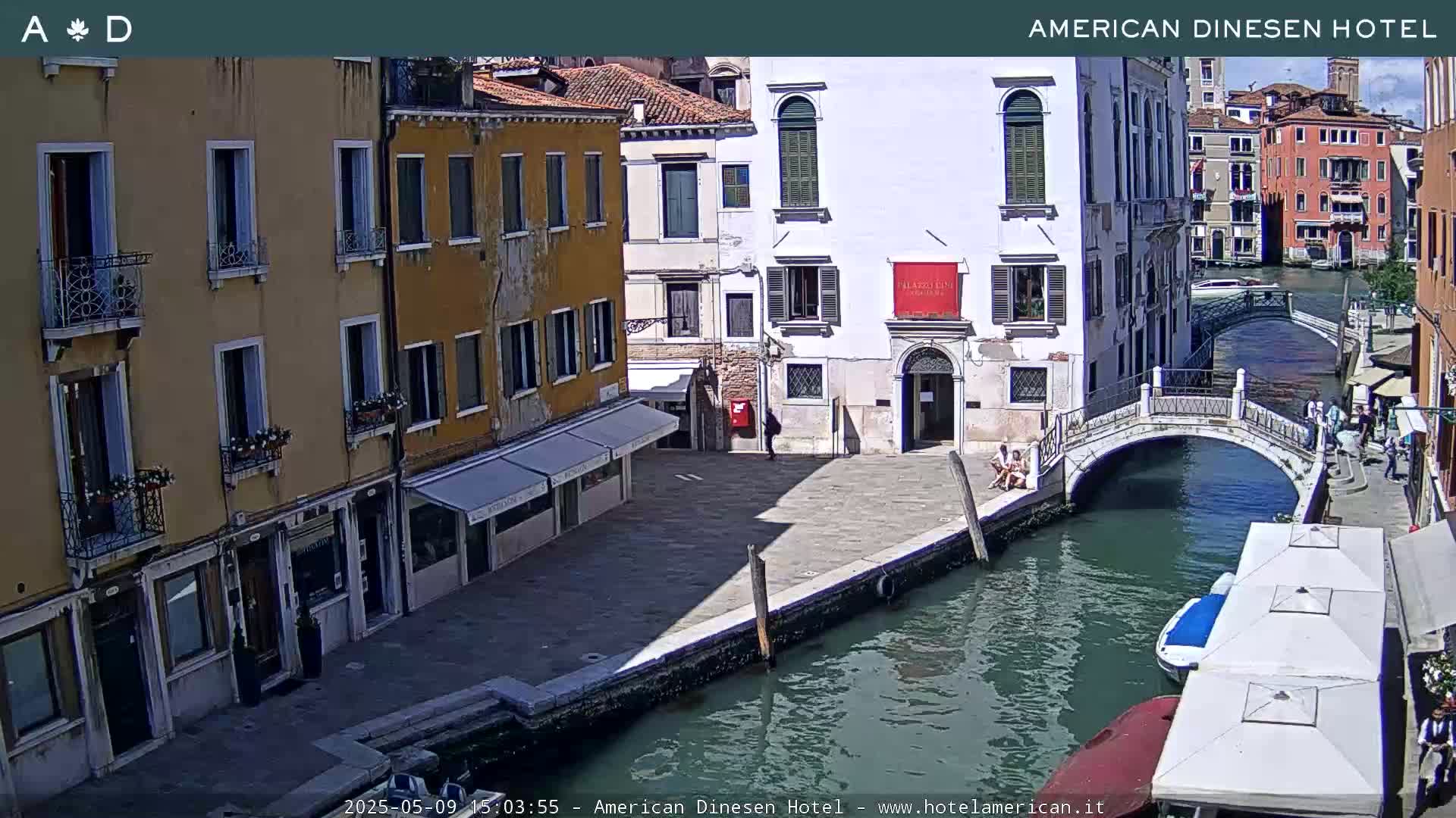 A sunny Venetian canal scene shows several buildings lining the waterway, a small bridge spanning the canal, and a few boats moored along the edge.