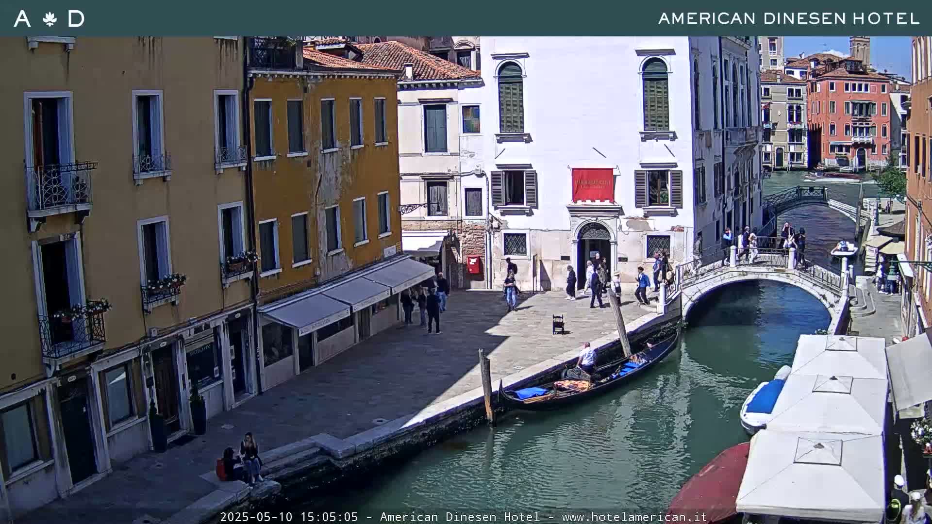 A sunny Venetian canal scene shows pedestrians walking along a canal-side walkway and across a small bridge, with gondolas in the water and buildings lining the canal.