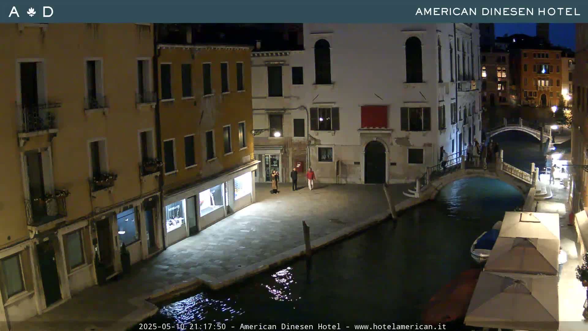 A nighttime view of a canal in Venice, showing several buildings along the water's edge, a small bridge, and a few people standing near the buildings.