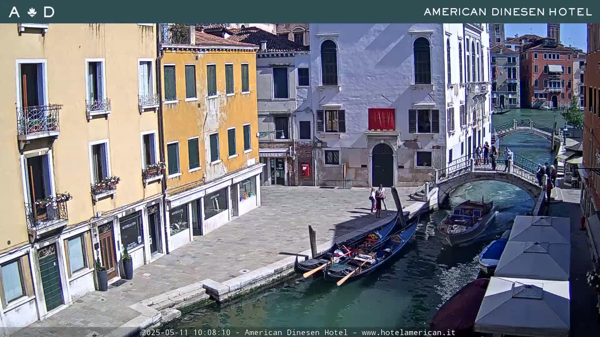 A sunny view of a Venetian canal with gondolas, pedestrians on a bridge, and colorful buildings lining the waterway.