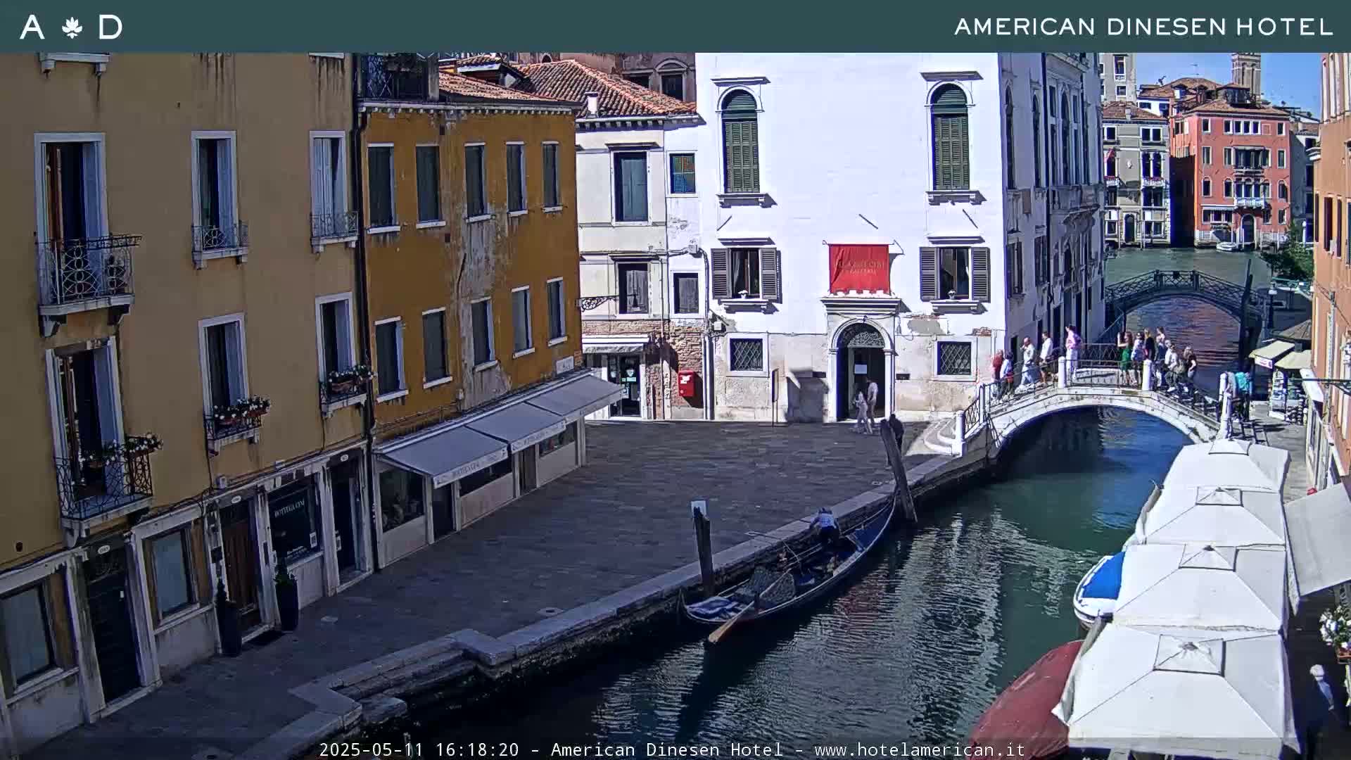 A sunny Venetian canal scene shows colorful buildings, a gondola, pedestrians on a bridge, and outdoor cafe umbrellas.
